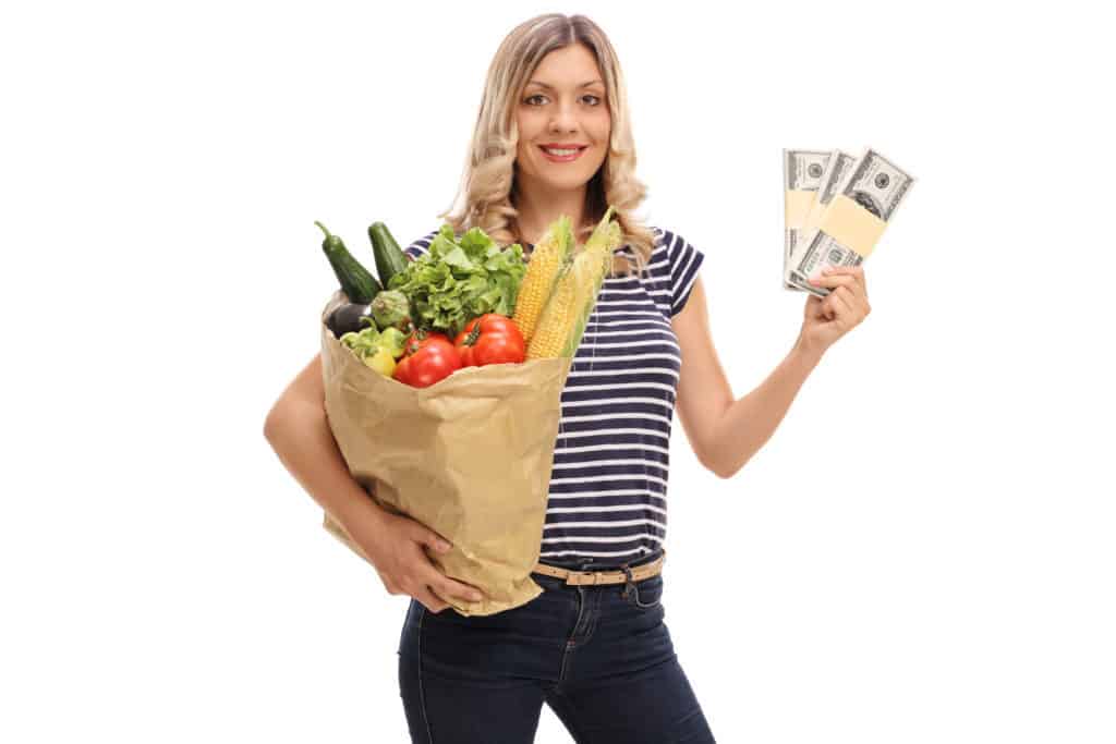 Smiling woman holding a bag of groceries in one hand and stacks of money in the other.